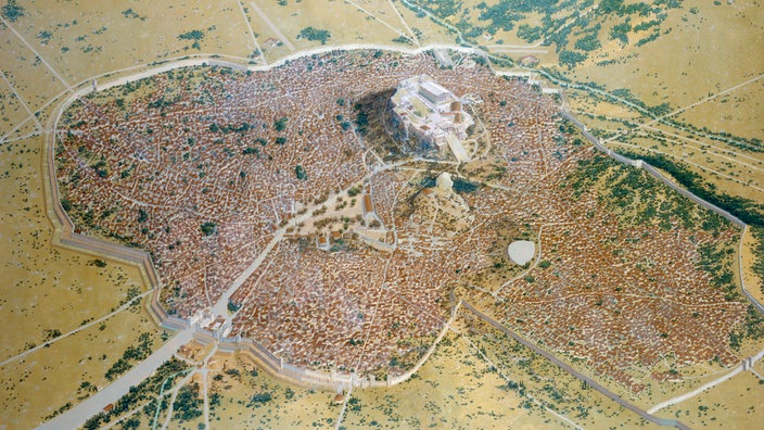 In einer Landschaft mit geringer Vegetation sieht man aus der Vogelperspektive eine Stadt liegen. Auffallend ist ein Hügel innerhalb der Stadt mit einer Tempelanlage.