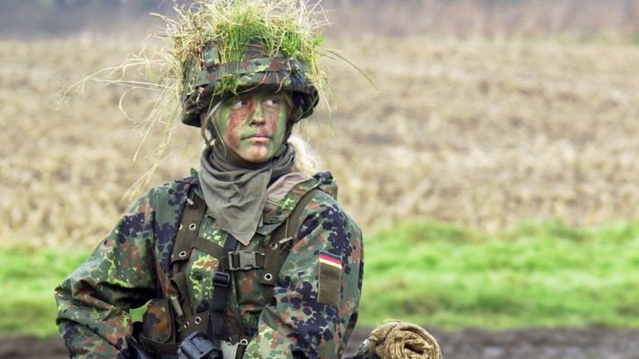 Mit der Waffe im Anschlag läuft eine junge Soldatin im Tarnanzug über eine Wiese.