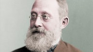 Ein koloriertes Foto aus dem Jahr 1887 zeigt den Mediziner und Reichstagsabgeordneten Rudolf Virchow. Er trägt einen dichten grauen Bart und eine Brille mit runden Gläsern. Bekleidet ist er unter anderem mit einer braunen Anzugjacke.