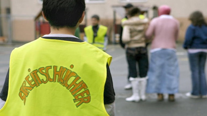 Ein Schüler in gelber Weste mit Aufschrift 'Streitschlichter' beobachtet den Schulhof während der Pause.