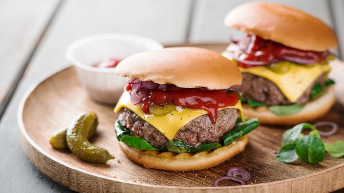 Zwei dick belegte Hamburger mit viel Ketchup auf einem Holzbrett. Dekorativ daneben eine Schale mit Ketchup, zwei Cornichons, einige Zwiebelringe und Basilikumblätter.