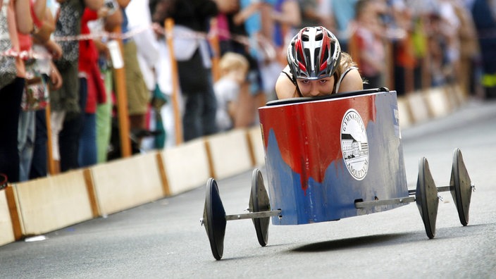 Eine Frau mit Helm fährt in einer Seifenkiste ein Gefälle hinab.