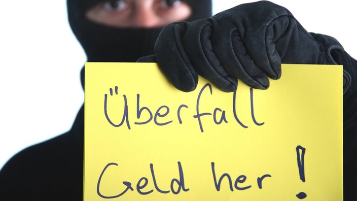 Vermummter Mann mit gelbem Schild mit der Aufschrift "Überfall Geld her!"