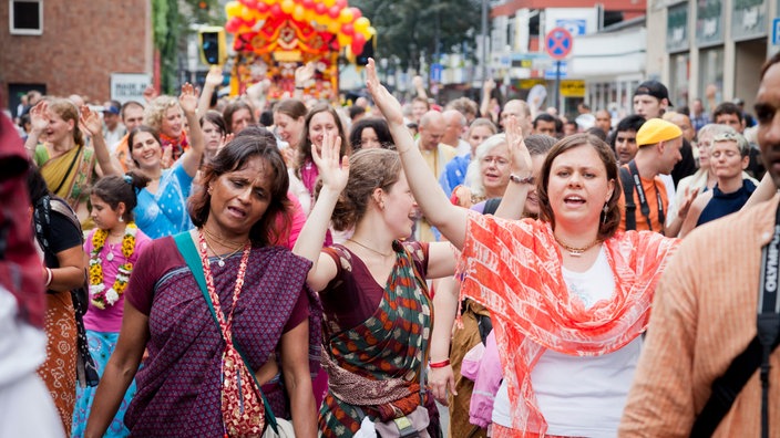 Viele bunt gekleidete Menschen gehen in einer Prozession singend durch die Straße