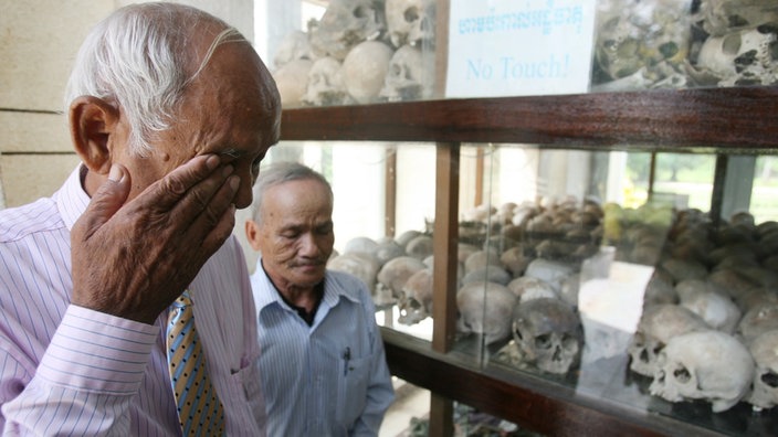 Zwei weißhaarige Männer stehen vor einer Vitrine mit Schädeln, einer reibt sich Tränen aus den Augen.