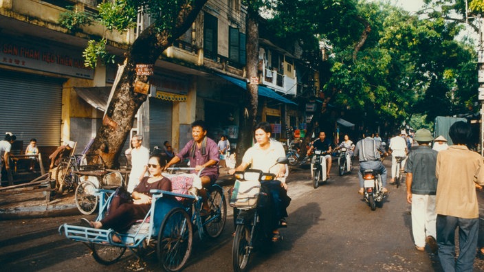 Straßenszene in Hanoi mit verschiedenen Fahrradfahrern und Fußgängern.