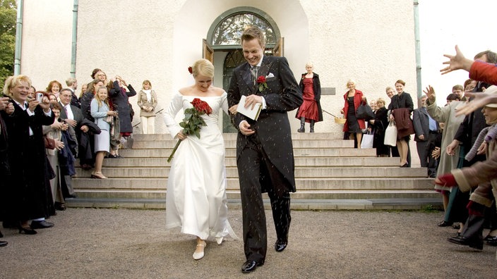 Ein Brautpaar schreitet vor einer Kirche die Stufen hinab. Rechts und links stehen die Hochzeitsgäste und werfen Reis.