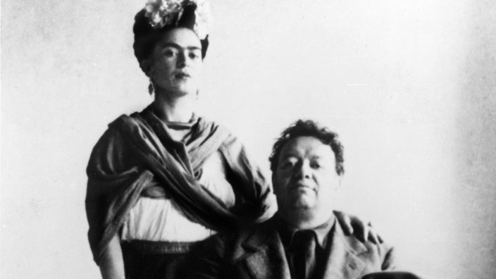 Schwarz-weiß-Fotografie von Frida Kahlo und ihrem Mann Diego Rivera; er sitzt auf einem Stuhl, seinen Hut in der Hand haltend. Sie steht hinter ihm und legt ihm eine Hand auf die Schulter.