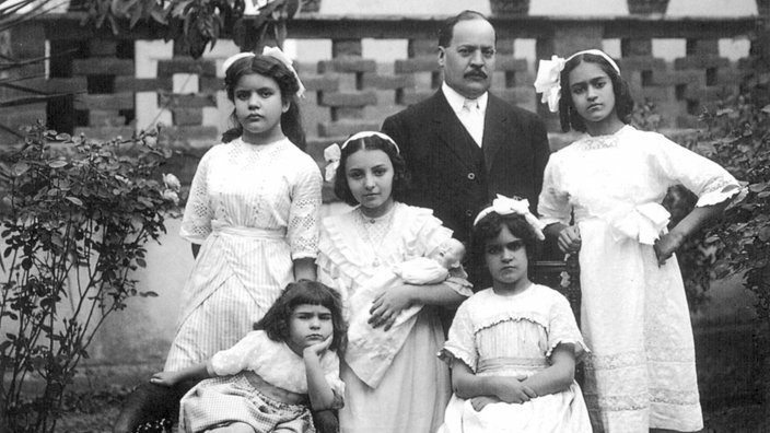 Schwarz-weiß-Fotografie der fünfjährigen Frida Kahlo mit ihren Schwestern Matilde und Adriana, zwei Cousinen und ihrem Onkel vor einer Mauer