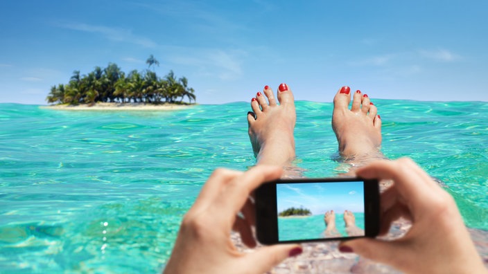 Eine Frau liegt im Meer und fotografiert ihre eigenen Füße, im Hintergrund eine Palmeninsel