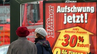 Zwei Frauen gehen an einem Werbeplakat vorbei