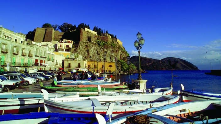 Blick über kleine Fischerboote auf den Hafen von Lipari mit Gebäuden an der Promenade. Im Hintergrund eine Festungsanlage auf einem vorspringenden Felsen.