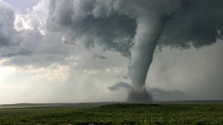 Flache Landschaft mit dunkel bewölktem Himmel aus dem ein Tornado zum Boden reicht.