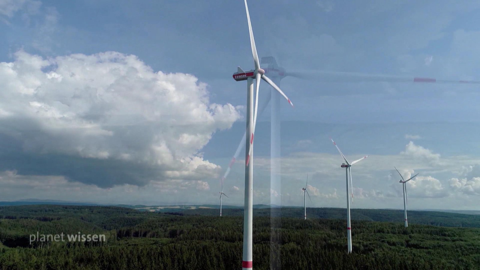 Mehrere Windkraftanlagen stehen hintereinander auf einem bewaldeten Höhenzug.