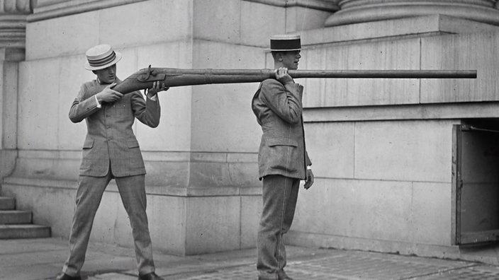 Schwarzweißfoto: Ein Mann mit einem riesigen Gewehr, dessen Lauf auf der Schulter eines zweiten Mannes aufliegt.