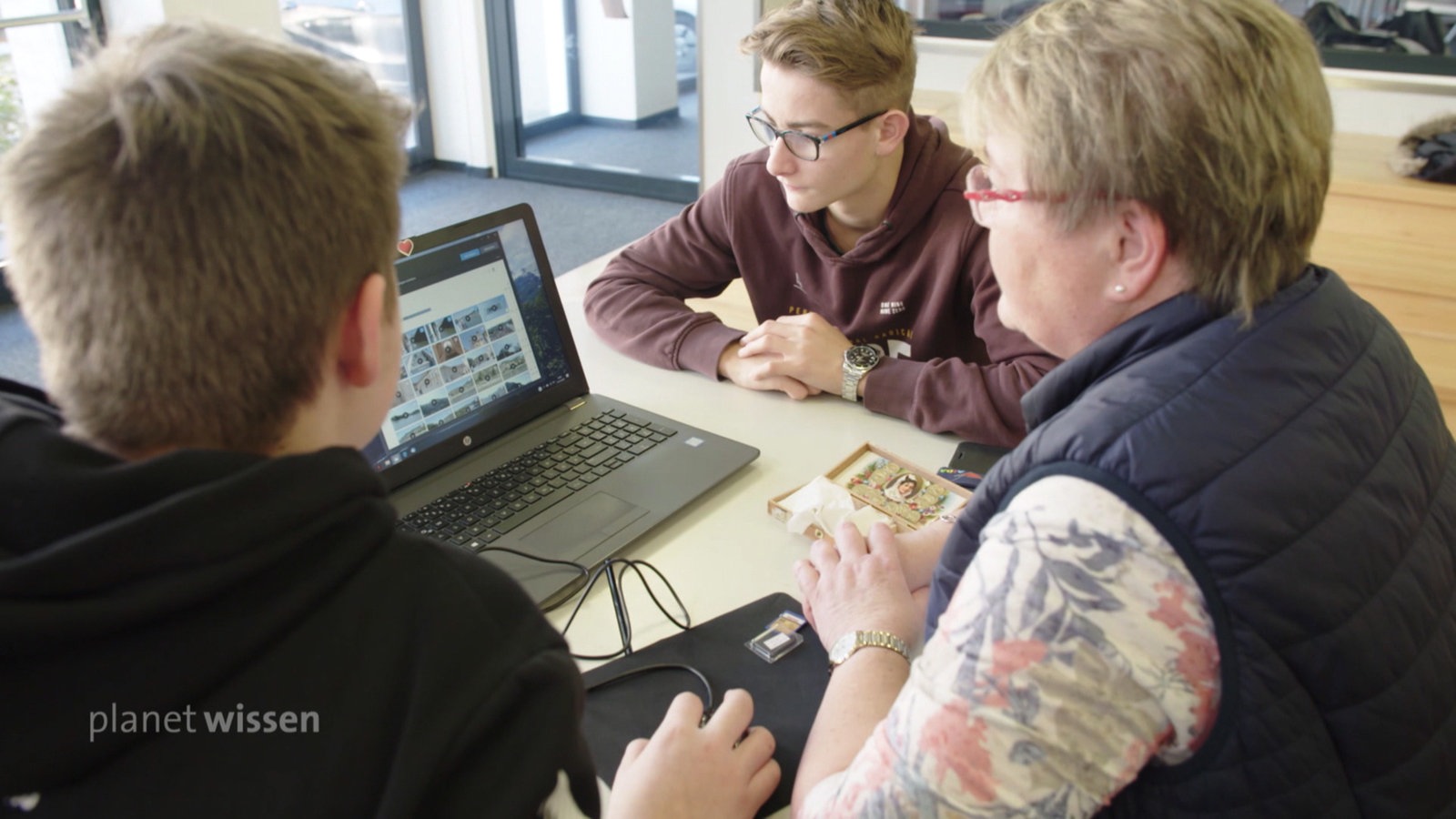 Eine ältere Dame sitzt flankiert von zwei jungen Teenagern vor einem Laptop.