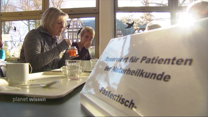 Zwei Frauen sitzen an einem Kantinentisch auf dem ein Schild steht 'Fastentisch'.