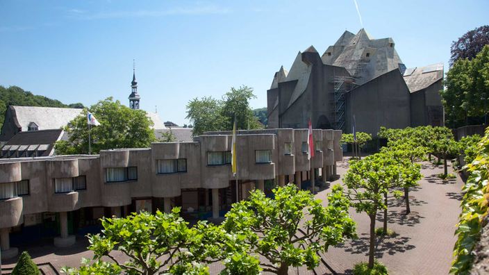 Moderne Kirche aus Beton mit einem Vorplatz, links kleine runde Häuser, die an Waben erinnern.